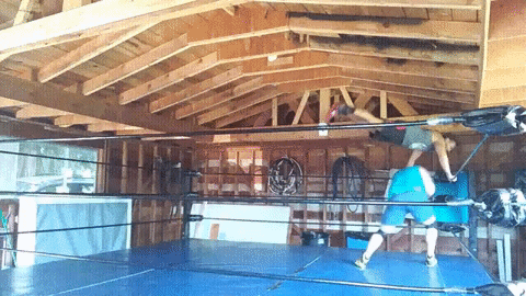 Two men in a wrestling ring performing a series of wrestling moves. One man jumps over the other in the corner, runs and bounces off the ropes, ducks a clothesline from the other man and shoulder tackles him to the ground