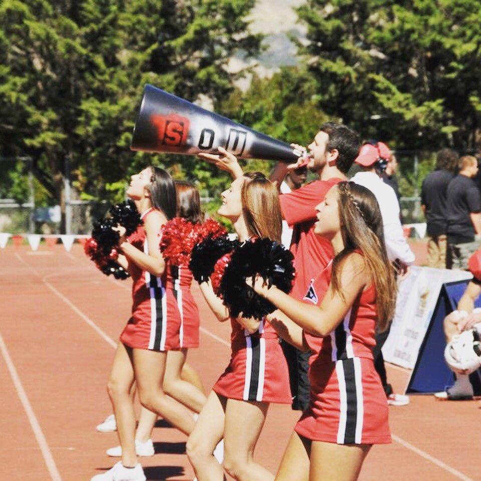 A group of femme cheerleaders in red  and black traditional cheerleading uniforms with red and black pom poms and a masculine cheerleader with a big black megaphone that has the letters "SOU" spray painted on it, are lined up on a running track performing a cheer.